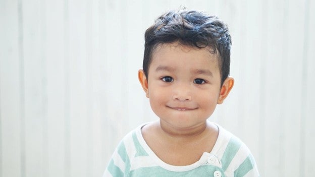 เด็กชายผมหยิก แก้มตุ่ย กำลังกัดริมฝีปากในชุดสีคาดลายฟ้าขาว